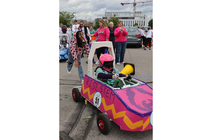 Push start. A student team pushes a handmade electric race car at the GEN‑EV competition as a helmeted driver sits inside, surrounded by other students, adults and spectators in a parking lot race course.