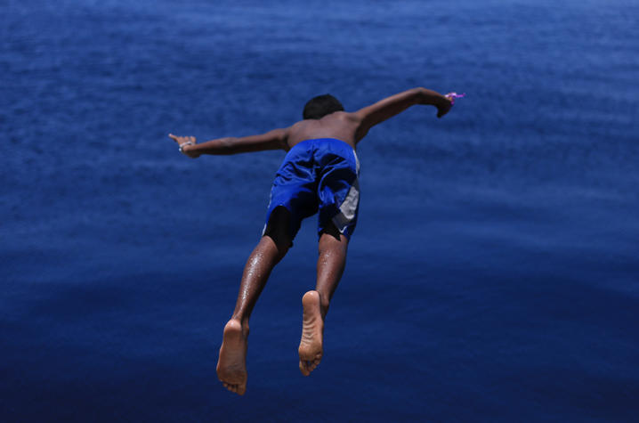 A child wearing blue shorts is captured midair in a horizontal position, arms outstretched, as they leap into deep blue water below.