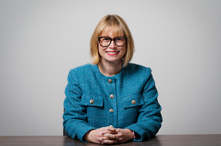 Studio portrait of a person wearing a blue textured jacket and dark-framed glasses, seated at a wooden table with hands folded, against a light gray background.