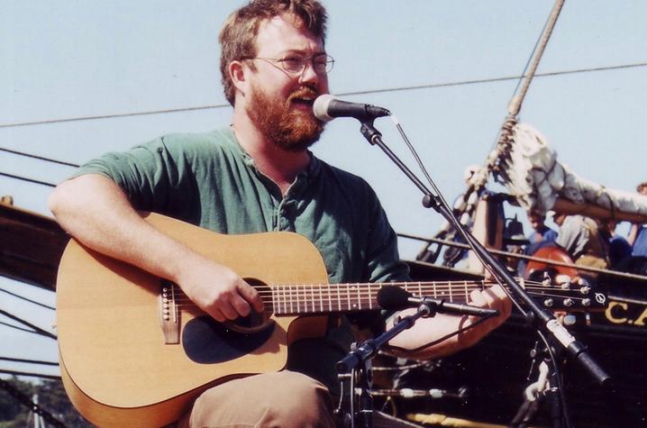 photo of Revell Carr with guitar at microphone singing sea chanteys in front of ship