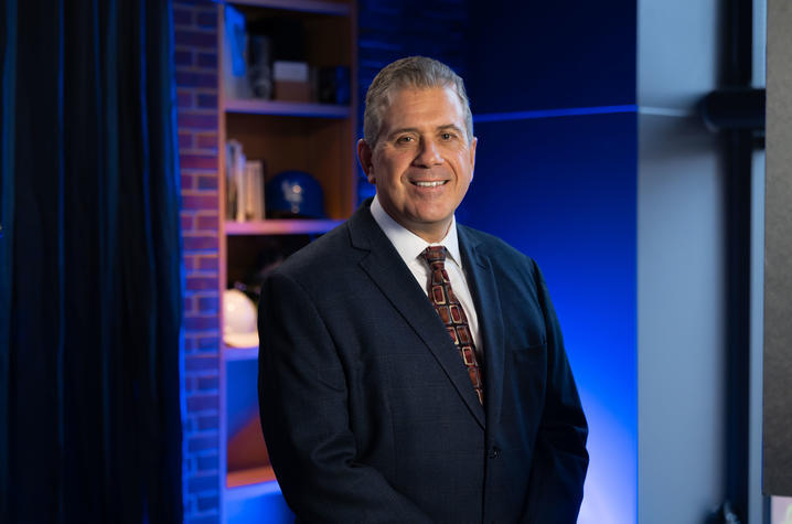 Person in a dark suit and tie standing in a studio with blue lighting.