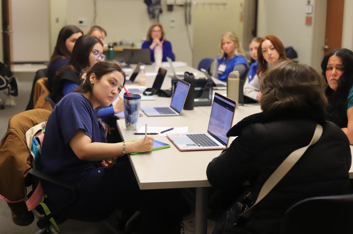 Health profession students sit around a conference table with laptops and tablets, taking notes and listening during a small-group training session.