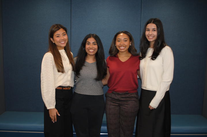 Four professionally dressed adults stand side by side indoors against a dark blue wall with a bench along it. They are close together with relaxed postures, suggesting a group or cohort photo taken in a workplace or professional program setting.