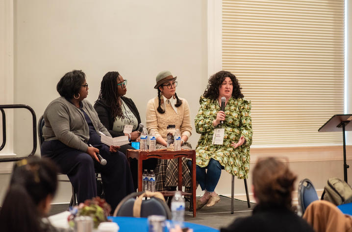 photo of presenters Four panelists sit on a stage during a conference discussion, with one panelist speaking into a handheld microphone.