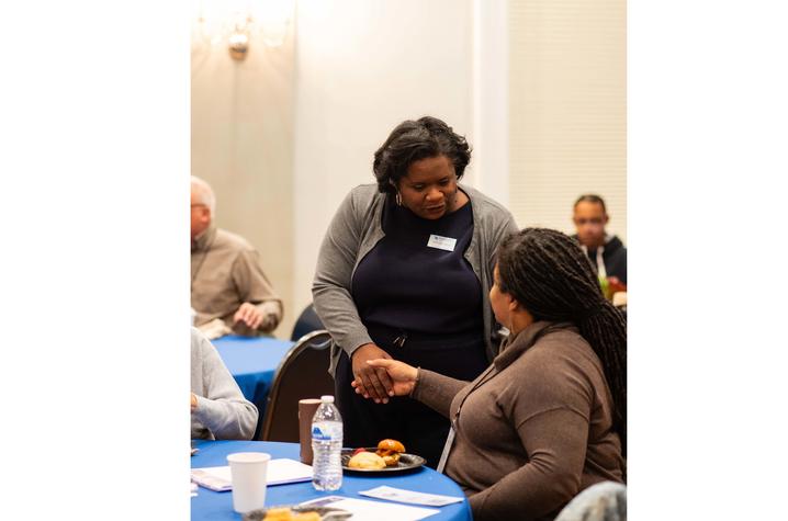 photo of Monuments Workshop co-founder Two attendees greet each other with a handshake during a networking or community event. The interaction takes place at a round table covered with a blue tablecloth, with notepads, a water bottle, and breakfast items visible.