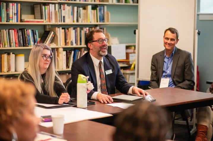 photo of Monuments Workshop co-founder Participants gather around a conference table during a small-group discussion in a library setting. Bookshelves filled with books line the background while attendees sit with notebooks and meeting materials.