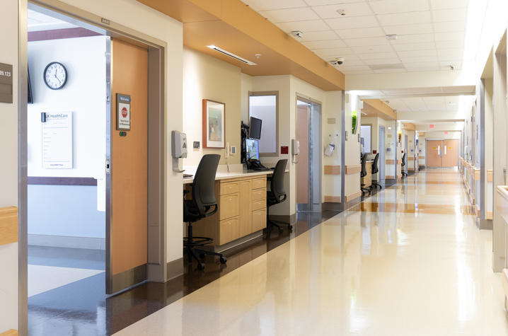 A bright hospital hallway with room doors along one side and a central nurses’ station with computers and chairs