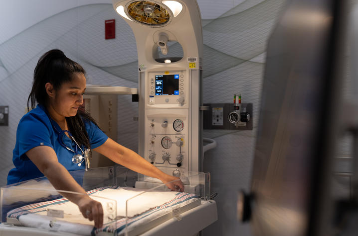A healthcare worker in scrubs adjusts equipment inside an infant warmer in a clinical room with medical monitors and controls