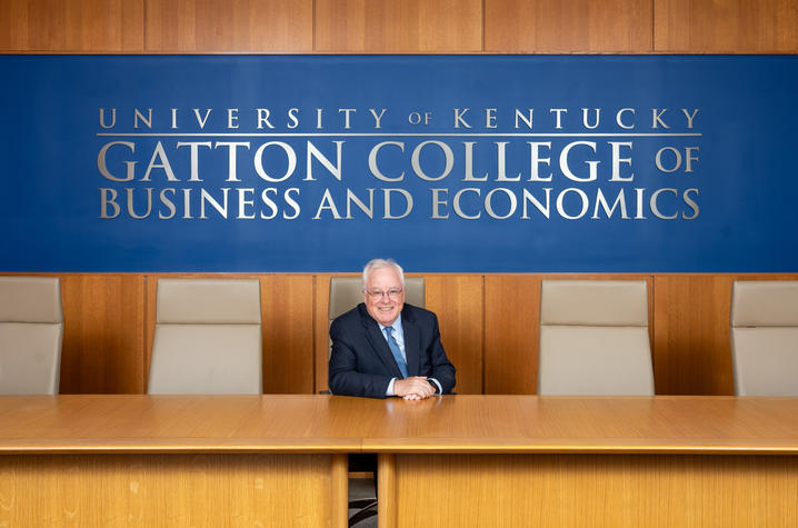 photo of Simon Sheather Person in a navy suit seated at a long wooden conference table beneath a blue wall sign reading “University of Kentucky Gatton College of Business and Economics,” with empty chairs on either side.