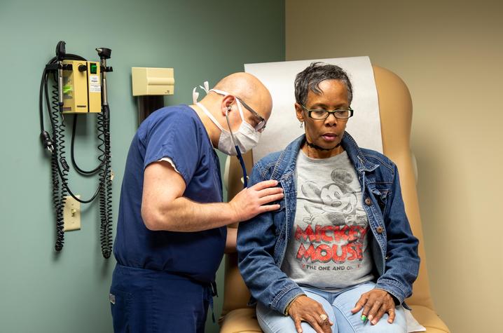 Image of doctor in blue scrubs listening to a woman's heart in a medical exam room