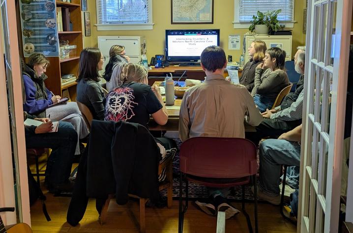 A small group of people sit in a cozy room watching a presentation on a TV screen.