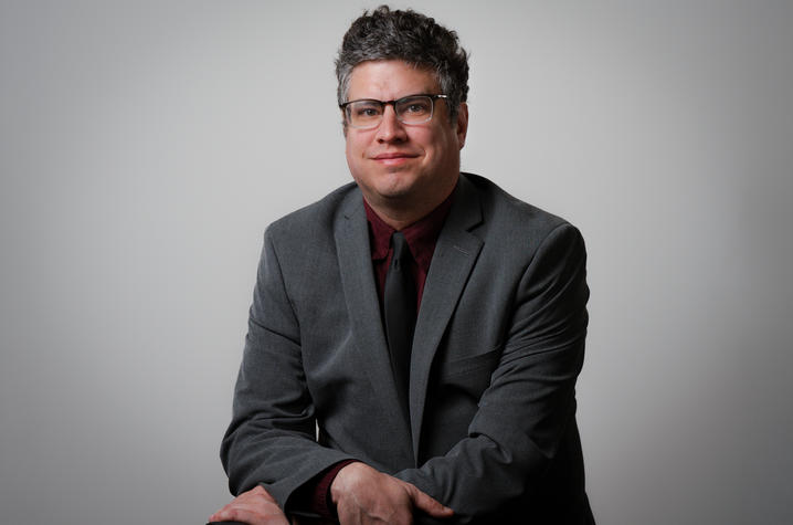 Scott Berry Studio portrait of a person with short curly hair and glasses, seated and smiling slightly. They wear a gray suit with a dark red shirt and black tie against a neutral light gray background.