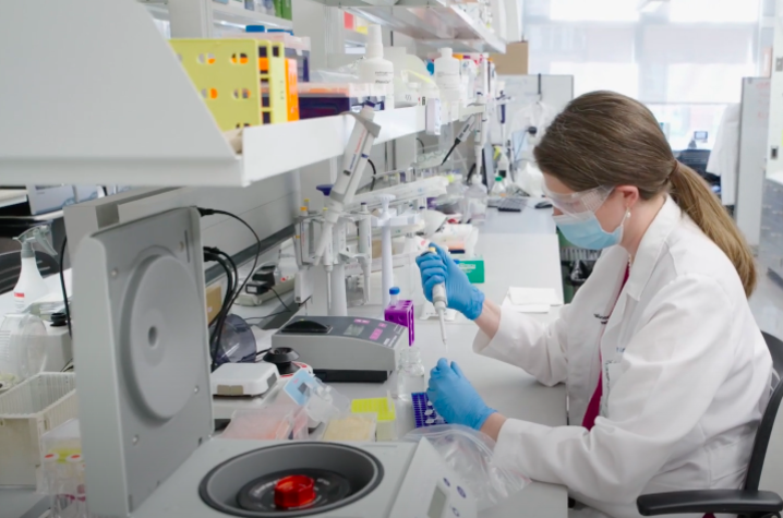 Profile photo of Dr. Mary Sheppard as sits at the workbench in her medical laboratory, using a pipette to put fluid into vials. She's wearing a white lab coat, blue medical gloves, goggles and a face mask. She's surrounded my medical lab equipment/tools. 