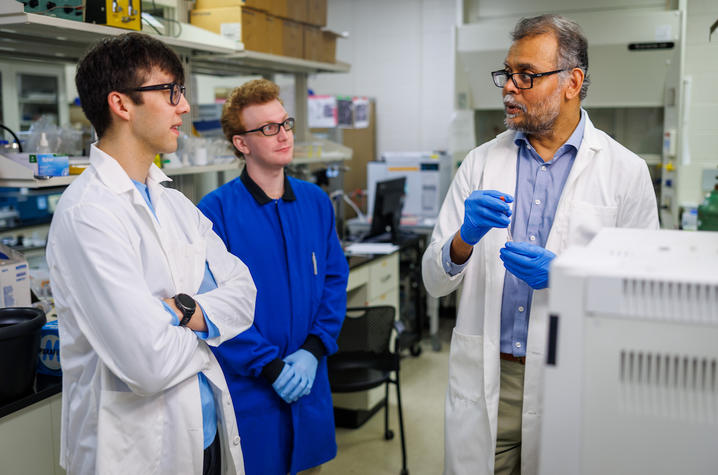 C. macellaria reearh team Three individuals wearing laboratory coats and protective gloves stand in a research laboratory beside benches and scientific equipment. One individual holds a small vial or tube while speaking to the other two, who are listening nearby.