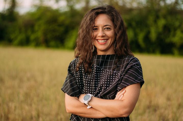 A person with long wavy hair stands in a grassy field with arms crossed, wearing a patterned black top and a smartwatch. Tall trees and greenery create a soft, natural backdrop under a lightly clouded sky.