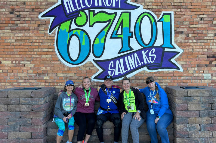 Brian Rice poses with the nurses who saved his life and his wife. Five people wearing race medals sit together on a stone ledge in front of a colorful mural that reads “Hello from 67401 Salina, KS.” They are smiling and posing with arms around one another.