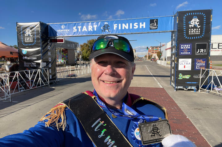 Brian Rice at the finish line A man wearing a blue Prince Charming-style costume takes a selfie while holding his marathon finisher medal. Behind him is the Crossroads Marathon start/finish archway on a sunny day.