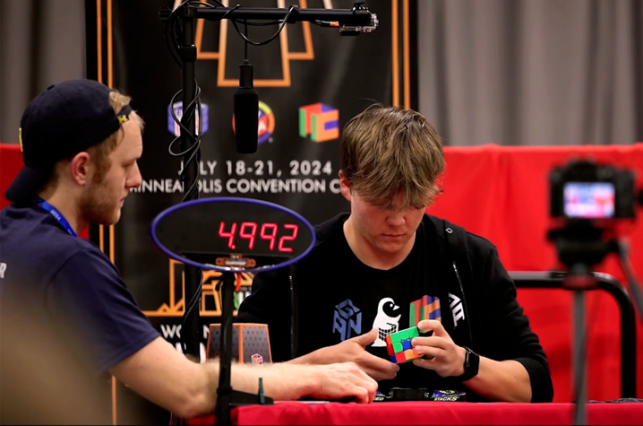 A competitor solves a Rubik’s Cube at a speedcubing event, with a timer showing 4.992 seconds and a camera recording the attempt.