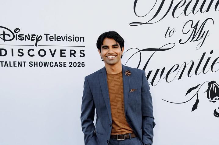 Person in a blue suit and brown shirt posing on a red-carpet event backdrop with Disney Television Discovers Talent Showcase branding.