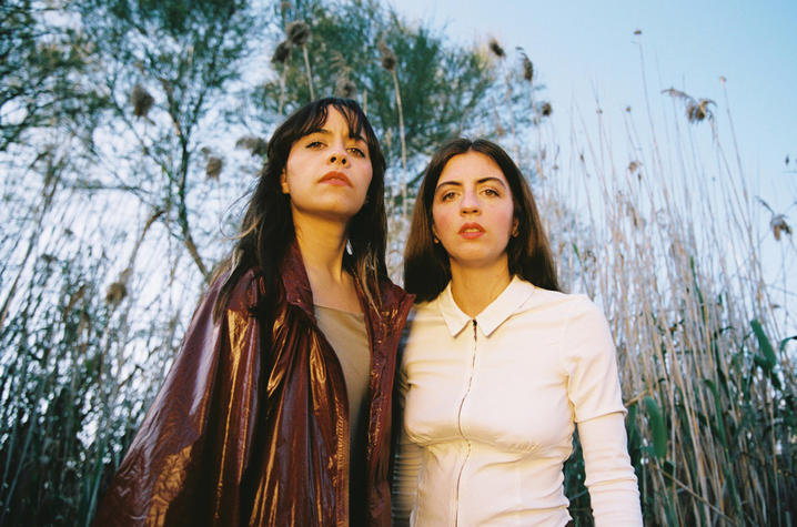 Two people standing outdoors among tall grasses and trees, wearing a brown jacket and a white top.