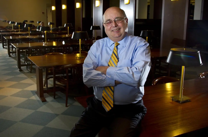 White male wearing yellow tie and blue shirt leaning on table in a library setting