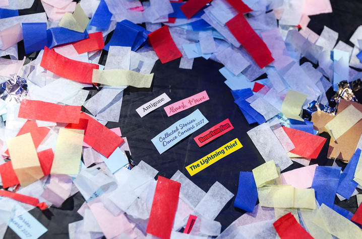 Colorful pieces of confetti in red, blue, white and pink surround a dark surface scattered with several small printed labels containing text.