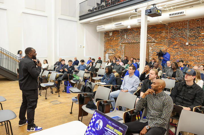 A speaker addresses an attentive audience in a lecture hall with exposed brick walls. The audience, seated in rows, listens intently, creating an engaging atmosphere.