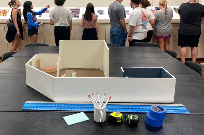 Model of an art gallery sitting on a table with a ruler, a cup of pens, tape and a tape measure, with students standing in the background looking at an exhibit with their backs to the camera