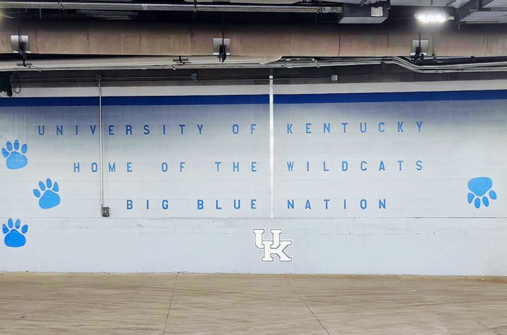 White wall inside a University of Kentucky loading dock painted with blue paw prints and the words “University of Kentucky Home of the Wildcats Big Blue Nation,” with a large UK logo centered near the floor.