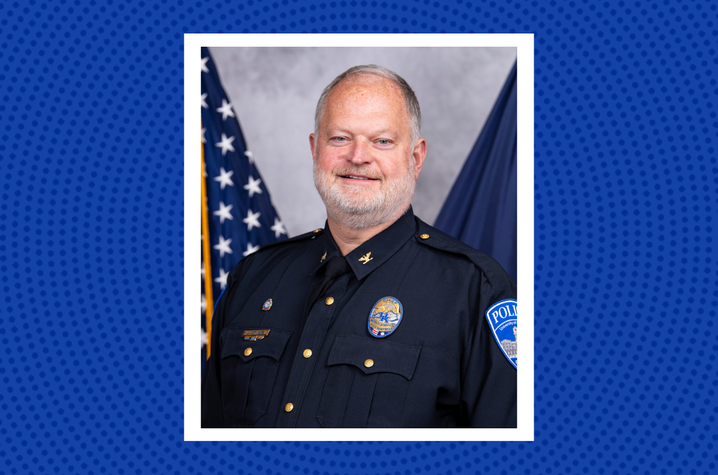 Photo of Chief Monroe elected as KACP president Police chief in a dark police uniform poses for a professional headshot. An American flag and a Kentucky state flag are visible in the background, with a blue patterned graphic border framing the portrait.