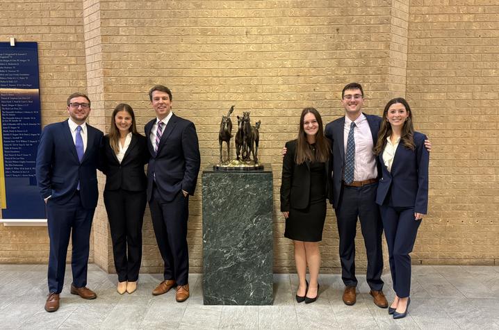 A group of six people dressed in professional business attire stand in a row inside a building with tan brick walls. They are positioned on either side of a green marble pedestal displaying a bronze horse‑themed sculpture.