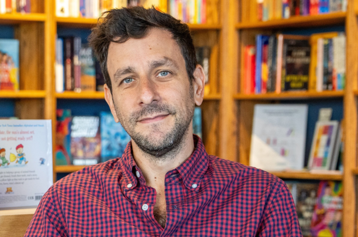 A person wearing a red and navy checkered shirt is seated in front of wooden bookshelves filled with colorful books. 