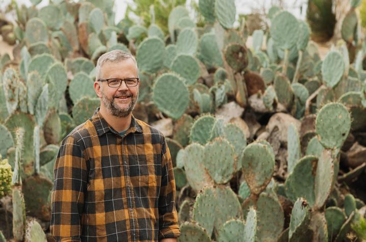 A smiling person wearing glasses and a brown plaid shirt stands outdoors in front of a dense cluster of green prickly pear cacti.