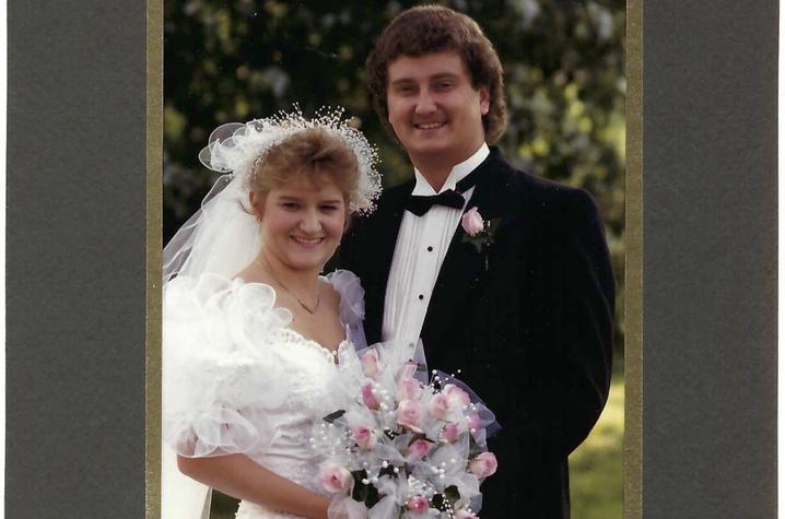 Wedding photo of young white couple. She's in a white dress with dramatic sleeves and a veil, holding a bouquet of pink roses. He's in a black tux and black bow tie. They're smiling at the camera, with trees behind them. 