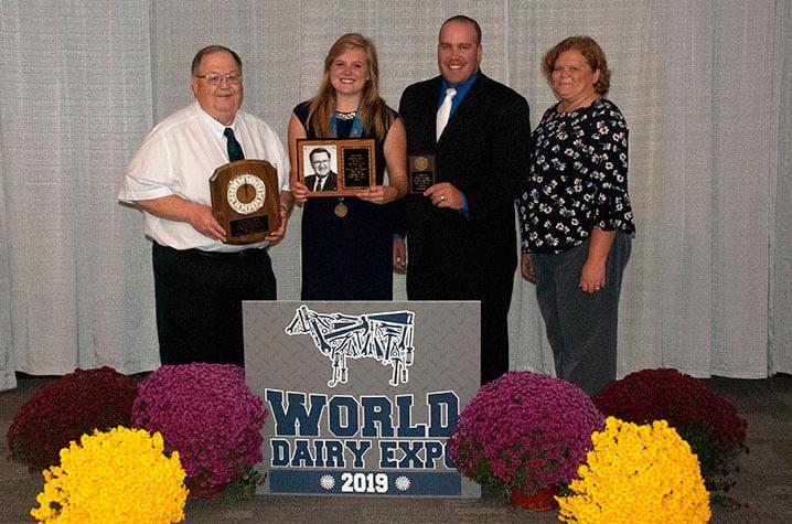 Four individuals stand on podium with awards and logo