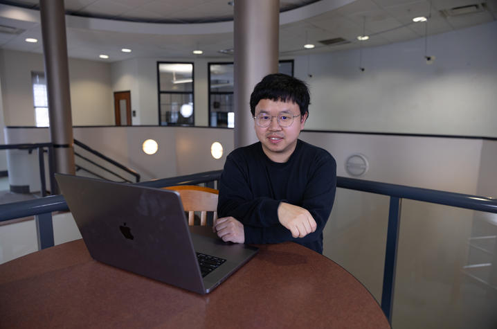 A person wearing a dark long‑sleeve shirt sits at a round table with an open laptop in front of them. 