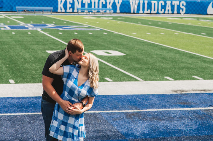 A couple stands on the edge of a football field, sharing a kiss. The woman wears a blue-and-white checkered dress, and the man wears a black shirt. Behind them, the stadium seating displays the words “Kentucky Wildcats,” and the field’s bright green turf 