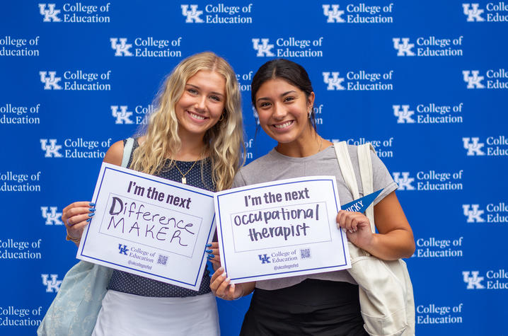 Two students standing in front of a UK College of Education backdrop holding signs that say I’m the next difference maker and I’m the next occupational therapist. 