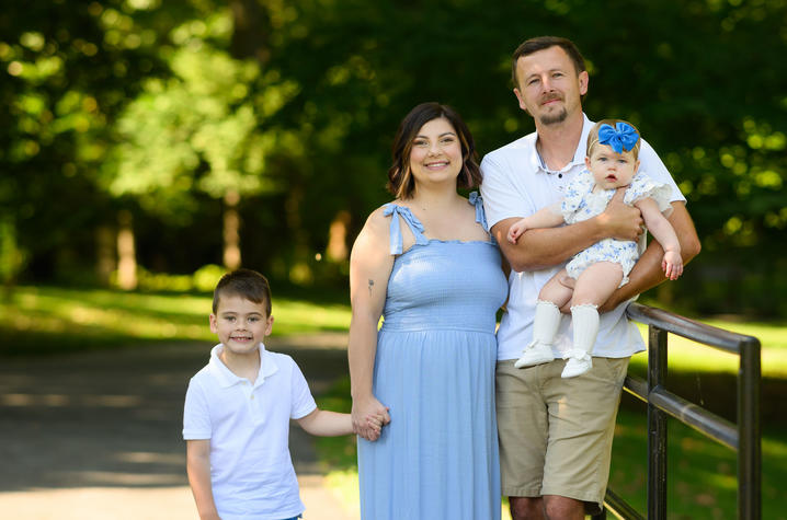 Image of the Wiseman family Family of four standing on a tree-lined park path, with one adult holding a baby wearing a blue bow while a young child stands beside them.