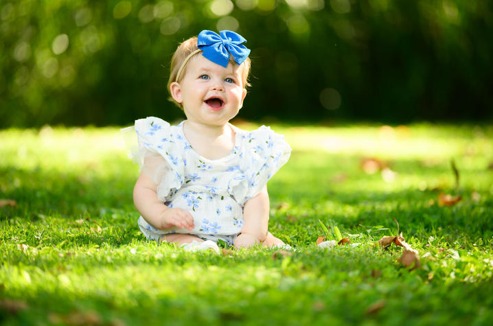 Image of Millie Wiseman A young child sits on green grass outdoors, wearing a white floral outfit and a large blue bow headband. The background is softly blurred with trees and sunlight filtering through the greenery.