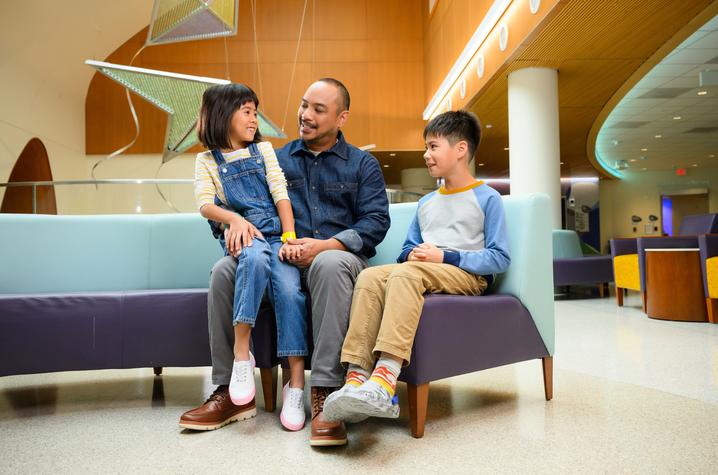 Family sits together on a curved bench in a bright hospital lobby, with one child seated on an adult’s lap and another child beside them. Modern star-shaped hanging art and warm wood accents are visible in the background.
