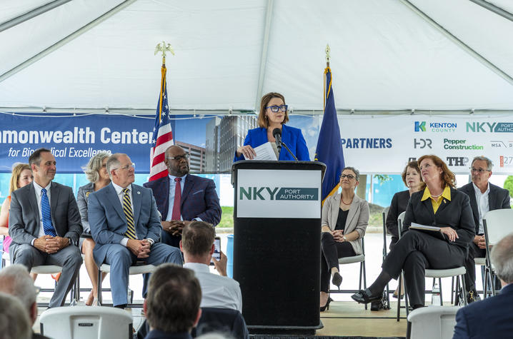 Photo of Holly Danneman A speaker stands at a podium under a tent during a groundbreaking ceremony for the Commonwealth Center for Biomedical Excellence in Covington, with seated officials, flags and event signage visible behind.