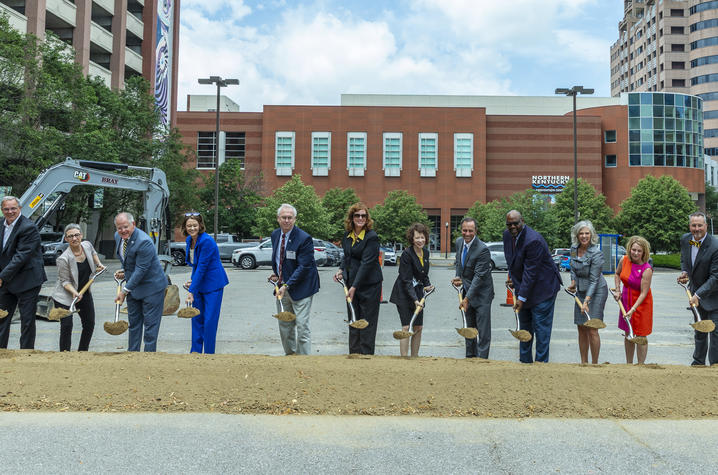 Photo of Commonwealth Center for Biomedical Excellence groundbreaking A group of about a dozen people stand in a line outdoors holding ceremonial shovels, participating in a groundbreaking event. The group is positioned on a patch of dirt and grass, with each person holding a shovel angled toward the ground.