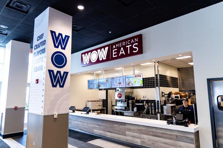 Interior view of a dining location labeled “WOW American Eats,” featuring a service counter, digital menu boards, beverage station, and a staff member working behind the register.