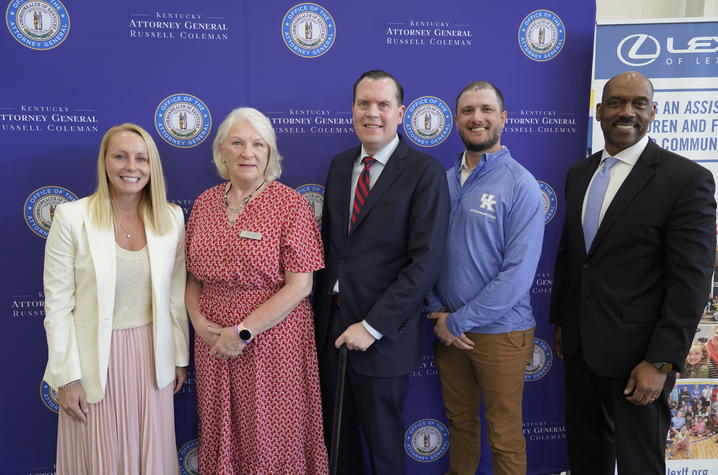 Five people stand in front of a blue Commonwealth of Kentucky-branded backdrop during an event, with a vertical display banner visible at the right side of the photo.