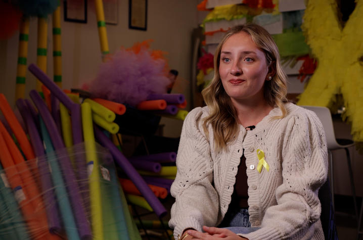 A person seated indoors during an interview, with colorful foam sticks and decorations in the background, wearing a yellow ribbon on a light sweater.