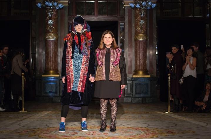 Two people standing on a patterned runway holding hands, wearing layered, colorful garments with intricate patterns in a grand hall.