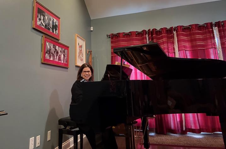 Person seated at a grand piano in a room with green walls, red curtains, and framed photos on the wall.