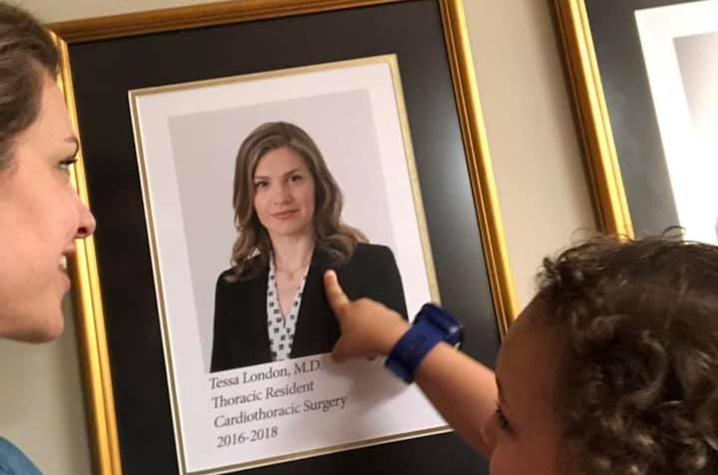 Child pointing at a framed portrait on a wall that includes text reading “Tessa London, MD, Thoracic Resident, Cardiothoracic Surgery, 2016–2018.”