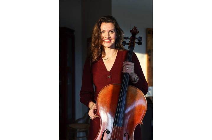 Person wearing a burgundy outfit holding a cello indoors with warm sunlight highlighting the instrument.